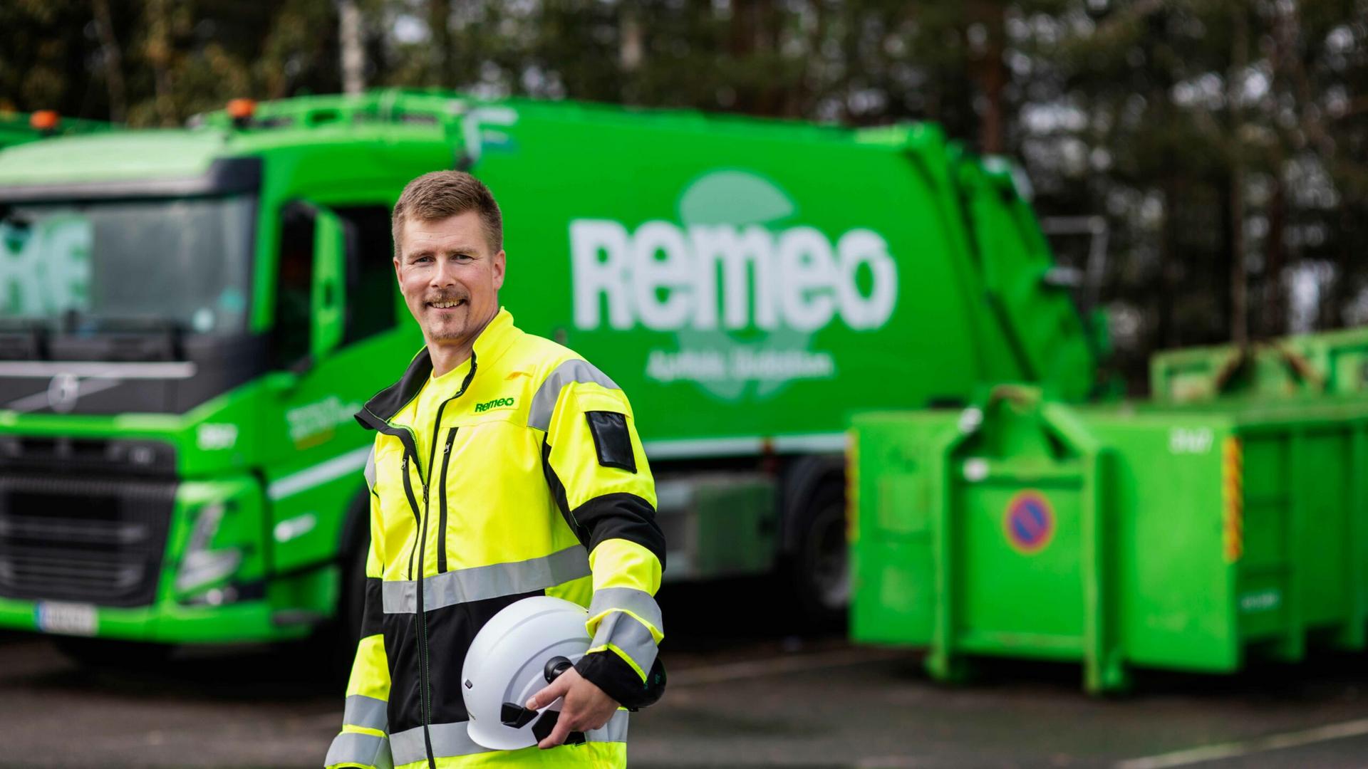 Remeon kuljettaja huomiovaatteissa katsoo hymyillen kameraan. Taustalla on Remeon vihreä jäteauto ja jätelava.